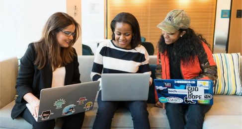 Three women sitting on a couch with their laptops, the two women on the left and right look onto the woman's laptop in the middle.