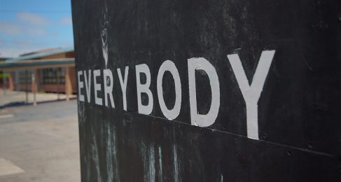 A photograph of a sign that reads the word everybody in white capitol letter on a black background. Off to the left appears to be a school structure but it is out of focus.