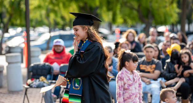Young fair skinned female with log reddish brown hair and a black cap and gown walks to collect her diploma.