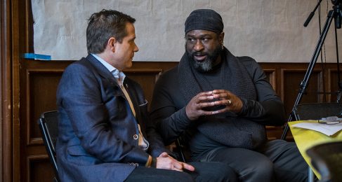 Two men, one of darker complexion and the other fair skinned are sitting down talking to each other at a conference. The darker complected man wears a black beanie and black suit jacket. The fair skinned man wears a blue shirt, yellow tie and a dark blue jacket.