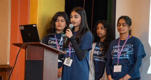 Four young women standing in front of a podium, three in the background and one on the forefront holding and speaking into a microphone. They all wear blue t-shirts that read "courage".