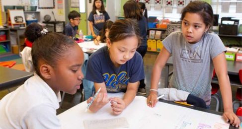 Three children sit and stand around a table, the girl inthe middle holds a pencil and all three are working on a project.